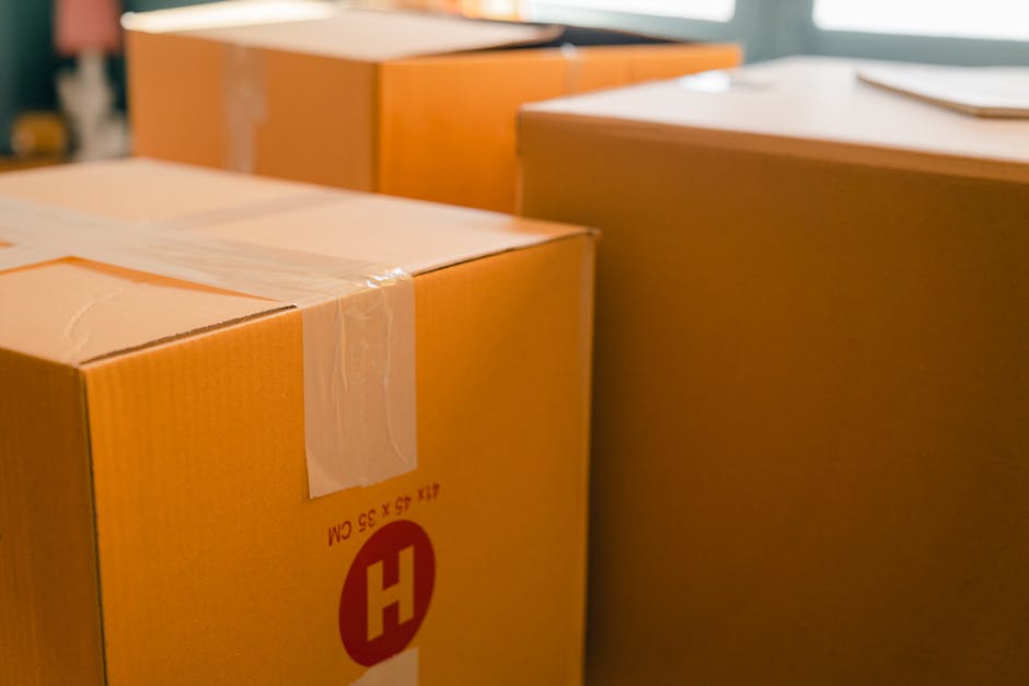 Close-up of several packed cardboard moving boxes with brown packing tape, stacked partially inside a home and near a doorway, ready for a home relocation. The boxes are situated on the floor near a wooden cabinet and a window, with natural light illuminating the scene. Some boxes display printed labels and weight indicators, reflecting careful packing and preparation for furniture transport. The environment suggests an interior space during a packing and moving process, with additional packaging materials visible in the background. This scene exemplifies the typical packing and loading stage in house removals, as handled by Man with Van Barking, with a focus on safe and organized relocation logistics, especially relevant during advice on avoiding parking fines during IG11 move days.