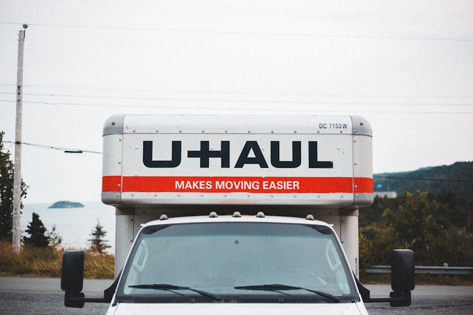 Front view of a U-Haul moving truck parked outdoors on a cloudy day, with the truck's cargo area visible at the top displaying the U-Haul logo and the slogan 'Makes Moving Easier' in red lettering. The windshield and side mirrors of the vehicle are clearly visible, with no driver or passengers present. The background features a rural landscape with a few trees, grassy areas, and distant hills under an overcast sky. This image captures the typical equipment used in home relocation, with the truck ready for transporting furniture, boxes, and appliances during packing and moving processes, and is associated with professional removals services like those provided by Man with Van Barking.