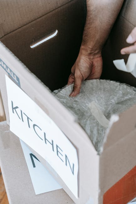 Close-up of several packed cardboard moving boxes with brown packing tape, stacked partially inside a home and near a doorway, ready for a home relocation. The boxes are situated on the floor near a wooden cabinet and a window, with natural light illuminating the scene. Some boxes display printed labels and weight indicators, reflecting careful packing and preparation for furniture transport. The environment suggests an interior space during a packing and moving process, with additional packaging materials visible in the background. This scene exemplifies the typical packing and loading stage in house removals, as handled by Man with Van Barking, with a focus on safe and organized relocation logistics, especially relevant during advice on avoiding parking fines during IG11 move days.