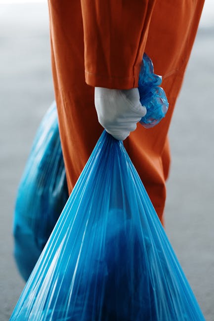 Close-up of a person wearing orange protective overalls and white gloves, holding a blue plastic rubbish bag filled with waste material. The individual is standing outdoors on a paved surface, with a blurred background. The scene reflects waste collection or disposal processes associated with home removals or moving activities, emphasizing proper handling and environmental considerations. This visual is relevant to house relocation services, such as those offered by Man with Van Barking, focusing on clearing or debris disposal during a move. The image highlights the importance of organized waste management as part of a professional furniture transport and packing process during home relocation.