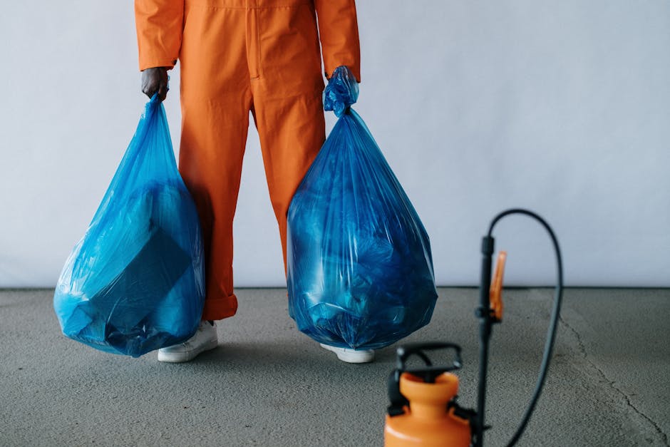 Close-up of a person wearing orange protective overalls and white gloves, holding a blue plastic rubbish bag filled with waste material. The individual is standing outdoors on a paved surface, with a blurred background. The scene reflects waste collection or disposal processes associated with home removals or moving activities, emphasizing proper handling and environmental considerations. This visual is relevant to house relocation services, such as those offered by Man with Van Barking, focusing on clearing or debris disposal during a move. The image highlights the importance of organized waste management as part of a professional furniture transport and packing process during home relocation.