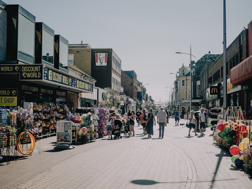 A wide view of a busy street in Barking with shops and market stalls on either side. The stalls display various items such as balloons, flowers, and toys, with some items placed in wire baskets and on tables covered with cloths. Several pedestrians are walking along the street, including a person pushing a stroller and others carrying bags, indicating a lively shopping area. The buildings housing the shops are multi-storied, featuring signage for brands like Discount World and McDonald's, along with other retail outlets. Street lamps are visible overhead, and the sky is clear, suggesting daytime with bright natural light illuminating the scene. The image captures a typical urban environment where home relocation and furniture transport services by Man with Van Barking could be relevant, especially during busy shopping hours or when coordinating moving logistics in a commercial area.