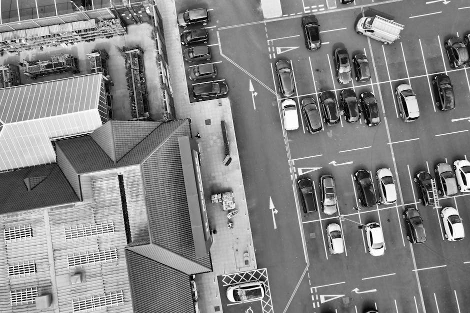 An aerial view of a parking lot adjacent to a building in Barking, showing numerous parked cars arranged in rows with white parking lines on asphalt. To the left, the building features a sloped roof with tiles, skylights, and a section with transparent roofing panels. There are concrete pathways alongside the building, with some stacked cardboard boxes and packing materials near the entrance. A man in a high-visibility vest is seen loading furniture or boxes onto a van that is parked close to the building, involving a lifting and carrying process. The parked van, part of the home relocation and moving services offered by Man with Van Barking, is positioned at the edge of the parking area, with the driver’s door open. The scene captures the logistics of packing and furniture transport during a house move, illustrating professional removal activities in an urban environment with clear daylight and no other moving vehicles in motion.