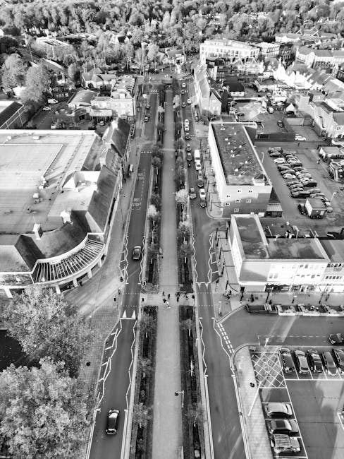 Aerial black-and-white photograph of a busy urban street in Barking Town Centre, showing the central road with vehicles parked along both sides and moving in both directions, divided by a median with trees and bushes. The street is flanked by commercial and residential buildings, some with flat roofs and others with pitched roofs, including a large modern building with a curved glass facade on the left and a smaller commercial structure on the right. In the foreground, a section of the pavement features pedestrian pathways marked with zebra crossings, and a few pedestrians are visible walking. The area also includes parking lots with cars neatly arranged. The photograph captures the overall layout of the street, highlighting the proximity of buildings, parking areas, and the main traffic flow, as part of the home or business relocation process that Man with Van Barking might facilitate for clients in the area.