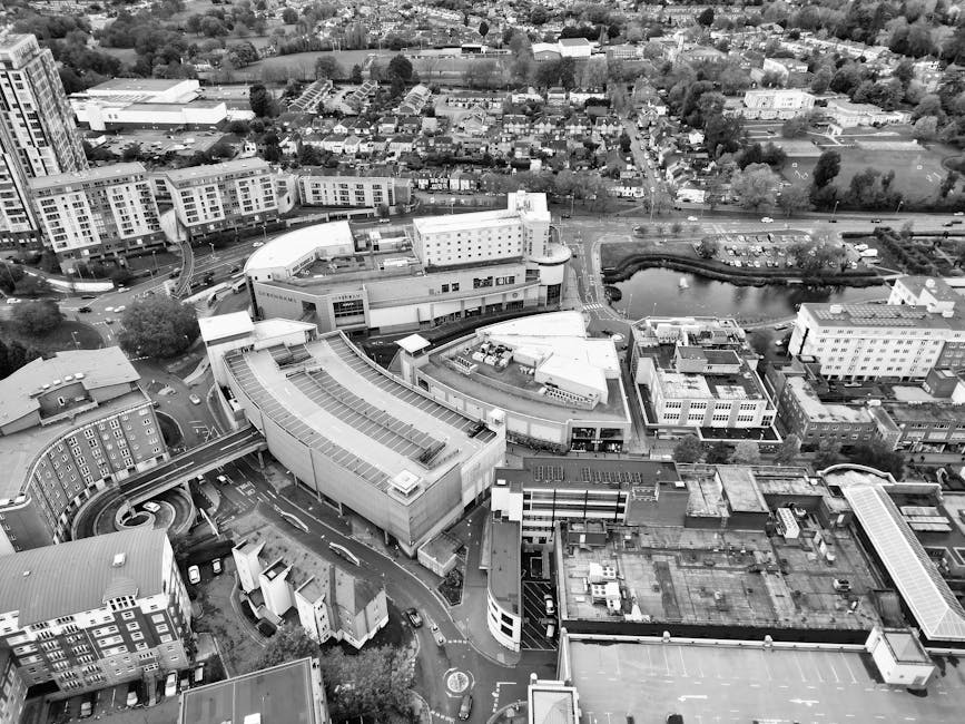 Aerial black-and-white photograph of a busy urban street in Barking Town Centre, showing the central road with vehicles parked along both sides and moving in both directions, divided by a median with trees and bushes. The street is flanked by commercial and residential buildings, some with flat roofs and others with pitched roofs, including a large modern building with a curved glass facade on the left and a smaller commercial structure on the right. In the foreground, a section of the pavement features pedestrian pathways marked with zebra crossings, and a few pedestrians are visible walking. The area also includes parking lots with cars neatly arranged. The photograph captures the overall layout of the street, highlighting the proximity of buildings, parking areas, and the main traffic flow, as part of the home or business relocation process that Man with Van Barking might facilitate for clients in the area.