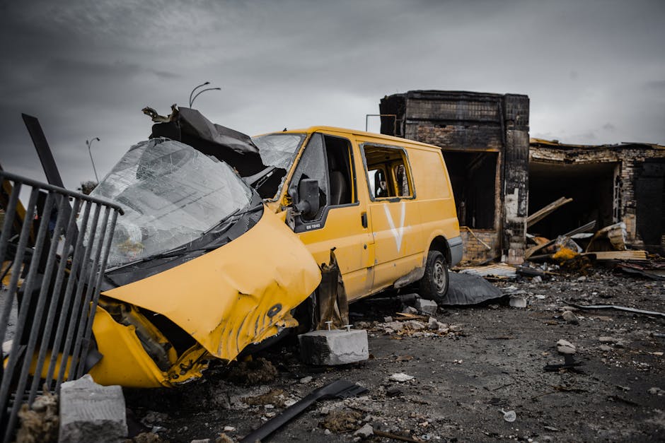 A yellow van with significant front-end damage and a shattered windshield is situated among debris and damaged structures, including broken bricks and charred building remnants, on a desolate, muddy g