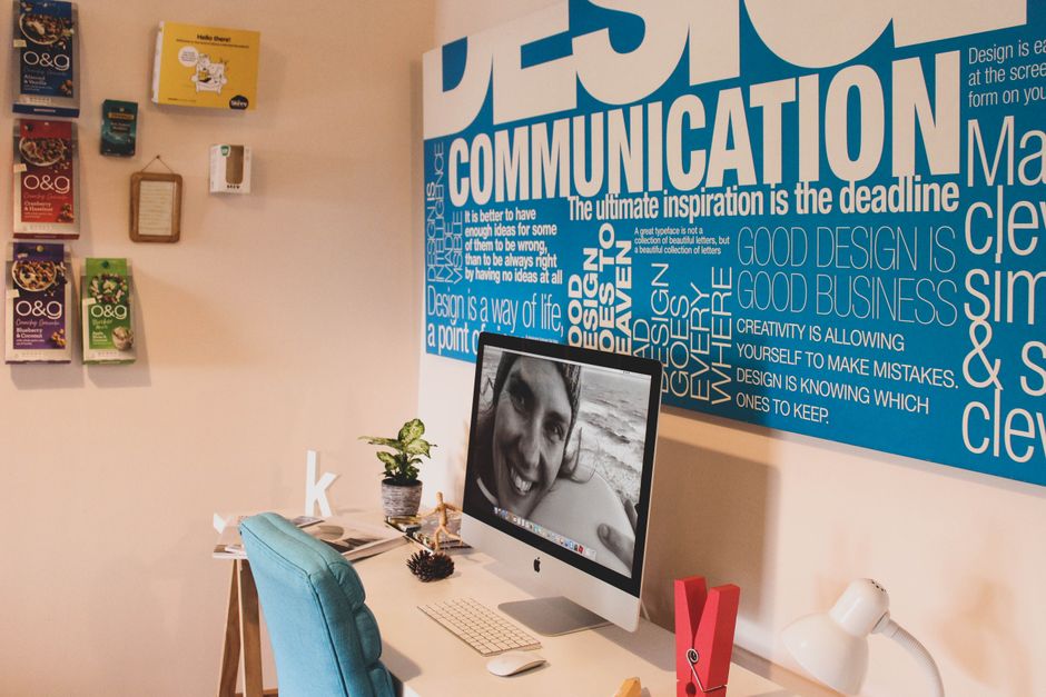 The image shows a home office interior with a white desk positioned near a light beige wall. On the desk, there is an all-in-one desktop computer displaying a black-and-white close-up photo of a smili