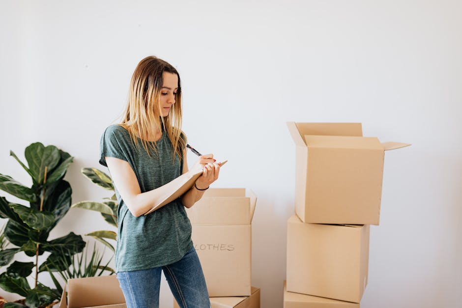 A young woman with long blonde hair, wearing a casual green t-shirt and blue jeans, stands inside a room with a plain white wall, surrounded by several packed cardboard boxes. She is holding a clipboard and writing with a pen, seemingly creating a moving inventory or checklist as part of a home relocation process. Behind her, there is a large green houseplant in a pot, and the boxes are stacked, some open and ready for packing or transport. The boxes, made of brown cardboard, are labeled with words like 'clothes' indicating packing categories. The setting suggests an organized packing or moving preparation, reflecting the type of logistics handled by [COMPANY_NAME] for furniture transport and removals. The scene captures the loading process or move planning, typical in house removals and packing services offered in Barking, and highlights the orderly approach to a successful move. The clear, natural lighting emphasizes the professional and practical environment typical for residential moving tasks.