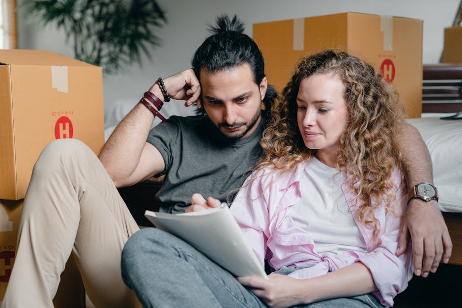 A man with dark hair tied back in a bun and a beard, wearing a grey t-shirt and beige trousers, is sitting on the floor amidst cardboard boxes and packing materials, with his arm resting on a box behind him. Beside him, a woman with curly blonde hair, dressed in a light pink shirt and grey trousers, is sitting on the floor holding a clipboard and pen, appearing to review a checklist. They are in an interior space that is well-lit, with several sealed cardboard boxes marked with red labels and tapes visible in the background, indicating a home relocation process. The scene depicts the packing and moving stage of house removals, with the pair focused on organizing belongings for transportation by a professional removals service such as Man with Van Barking. The environment suggests a typical room prepared for a house move, with the two individuals possibly reviewing a legal checklist for tenants moving out, as referenced in the page title, within a professional setting related to furniture transport and moving logistics.