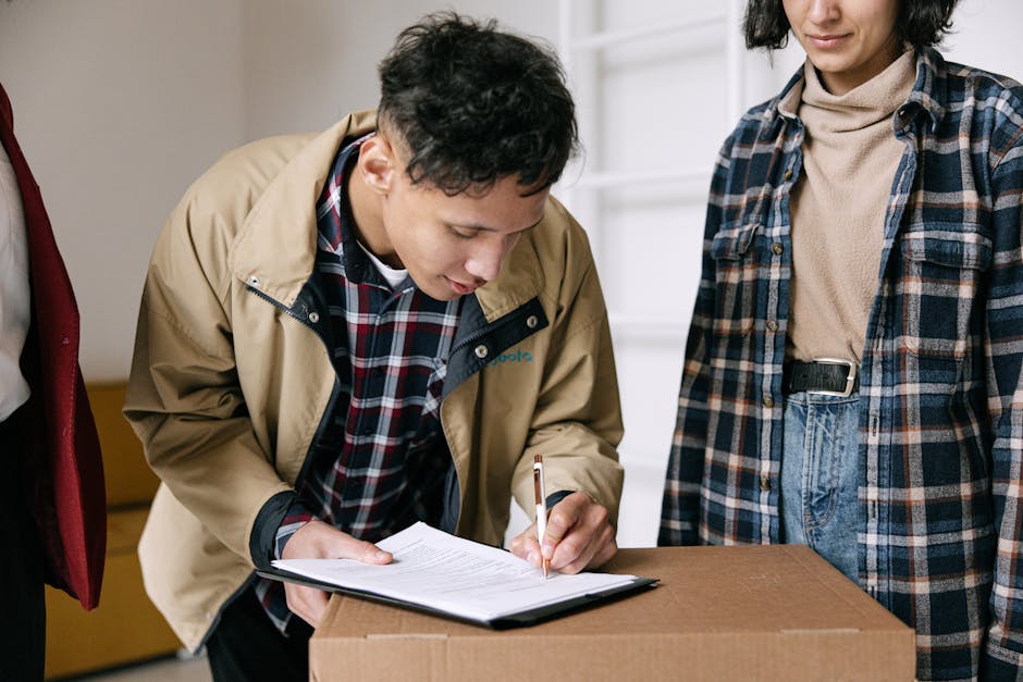 A young woman with long blonde hair, wearing a casual green t-shirt and blue jeans, stands inside a room with a plain white wall, surrounded by several packed cardboard boxes. She is holding a clipboard and writing with a pen, seemingly creating a moving inventory or checklist as part of a home relocation process. Behind her, there is a large green houseplant in a pot, and the boxes are stacked, some open and ready for packing or transport. The boxes, made of brown cardboard, are labeled with words like 'clothes' indicating packing categories. The setting suggests an organized packing or moving preparation, reflecting the type of logistics handled by [COMPANY_NAME] for furniture transport and removals. The scene captures the loading process or move planning, typical in house removals and packing services offered in Barking, and highlights the orderly approach to a successful move. The clear, natural lighting emphasizes the professional and practical environment typical for residential moving tasks.