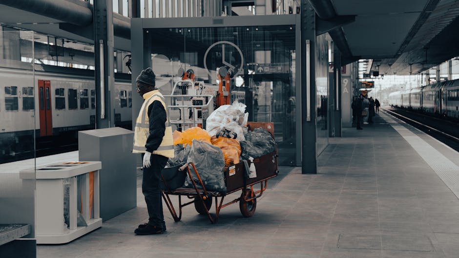 A man dressed in a dark jacket, grey beanie, and white gloves is standing next to a red trolley loaded with various bulky waste items, including black, orange, and grey plastic bags, cardboard boxes, and pieces of fabric. The trolley is positioned on a paved area of an outdoor train station platform with modern glass and metal structures, and a glass shelter behind the man. In the background, there are train tracks and a train with a white and grey exterior, along with a few other commuters visible in the distance. The scene is illuminated by natural light, indicating daytime, and the environment suggests preparation for waste disposal or transportation as part of a home relocation or moving process, which Man with Van Barking occasionally supports within their removals services.