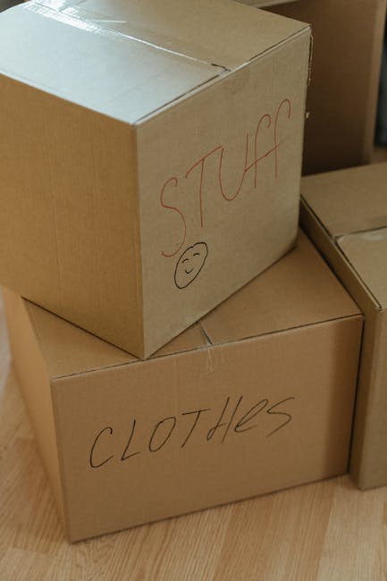 Indoor scene showing two medium-sized cardboard boxes stacked on top of each other, situated on a wooden floor. The top box has handwritten text in red marker reading 'STUFF' with a small smiley face drawn below it. The bottom box is labeled 'CLOTHES,' also handwritten in red marker. Both boxes are sealed with packing tape, and additional cardboard boxes are visible nearby, suggesting a packing and moving process within a home. This setting reflects typical home relocation activities, with the boxes prepared for loading into a van, and is associated with professional removals services such as those provided by Man with Van Barking, especially in the context of house removals or furniture transport for a property in Barking Riverside.
