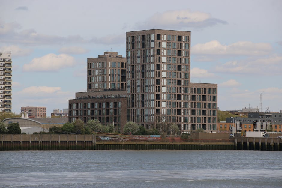 A modern multi-story residential building with a staggered, stepped design situated along the waterfront, featuring numerous rectangular windows and dark brick exterior cladding. The building is positioned behind a wooden barrier that lines the edge of the water, with some trees and shrubs visible at ground level. In the foreground, the water surface is calm, reflecting a partly cloudy sky with white and gray clouds. The surrounding area includes other low-rise buildings and distant structures, all part of an urban landscape. This scene relates to home relocation or furniture transport, as it visually depicts an urban setting where residents may engage Man with Van Barking for house removals and packing and moving services.