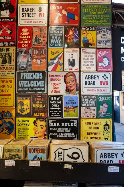Close-up view of a display wall covered with various colourful, vintage-style metal signs and stickers featuring humorous sayings, advertisements, and icons related to London, Ireland, and automotive themes. Beneath the signs, a wooden shelf holds a stack of cardboard boxes and a small portion of a box featuring the 'Abbey Road' design. The scene is set outdoors on a pavement with a black metal barrier in the foreground, and the signage appears to be part of a used goods or souvenir stall. This image, associated with home relocation and moving logistics, showcases the type of items that might be packed or transported during a house move, with specialist local removals company, Man with Van Barking, involved in such furniture transport and packing and moving services.