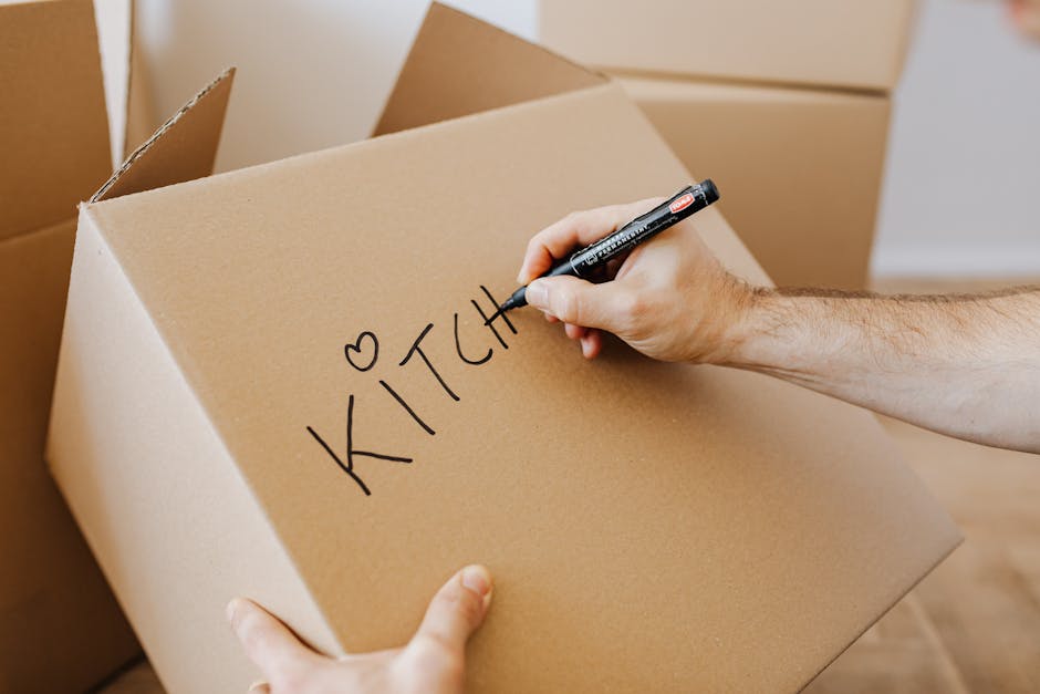 A person is holding a large cardboard box labeled 'KITCHEN' with a black marker, preparing it for packed items as part of a home relocation process. The individual is inside a room with several similarly sized cardboard boxes, some open and some closed, indicating the stages of packing kitchenware and utensils. The cardboard boxes are stacked on the floor, and the person’s right hand is writing the label while their left hand steadies the box. The scene shows a well-lit environment, likely a room in a house or flat, with the focus on the packaging process for moving. This image exemplifies the packing and moving preparation undertaken by [COMPANY_NAME], such as Man with Van Barking, during a house removal or furniture transport, emphasizing the importance of organized packing in residential relocations.