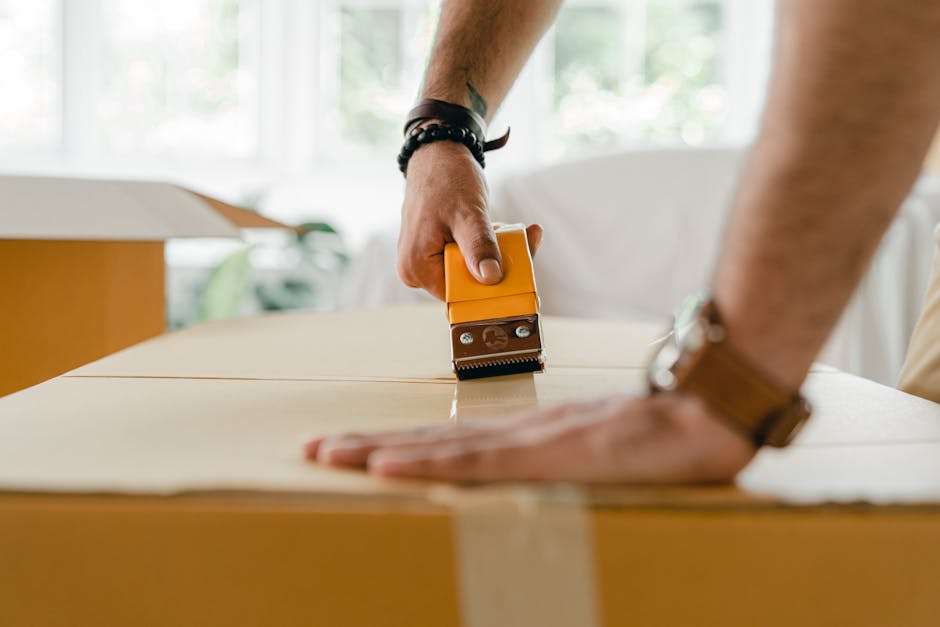 A person is using a box cutter or utility knife to carefully cut along a brown cardboard box, which is positioned on a flat surface inside a well-lit room with large windows in the background. The individual’s left hand is steadying the box while the right hand holds the cutter. The person is wearing a watch on their left wrist and a leather bracelet on their right wrist, suggesting a casual but practical approach to packing and moving. The scene captures the detailed process of preparing household items for a home relocation, consistent with services offered by Man with Van Barking, including careful packing for furniture transport and moving logistics. The process is part of the packing and moving stage of a house removal, with the focus on securing belongings in quality packaging materials such as cardboard boxes, which are essential for safe transport during house moves in the Gascoigne Estate area of Barking.