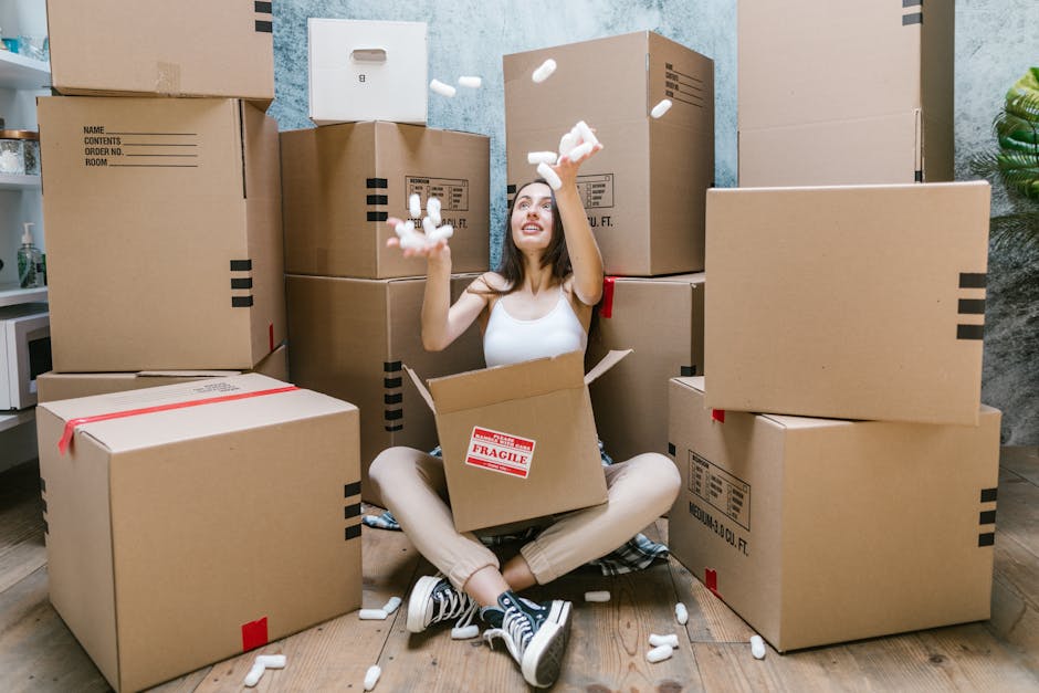 A person is holding a large cardboard box labeled 'KITCHEN' with a black marker, preparing it for packed items as part of a home relocation process. The individual is inside a room with several similarly sized cardboard boxes, some open and some closed, indicating the stages of packing kitchenware and utensils. The cardboard boxes are stacked on the floor, and the person’s right hand is writing the label while their left hand steadies the box. The scene shows a well-lit environment, likely a room in a house or flat, with the focus on the packaging process for moving. This image exemplifies the packing and moving preparation undertaken by [COMPANY_NAME], such as Man with Van Barking, during a house removal or furniture transport, emphasizing the importance of organized packing in residential relocations.